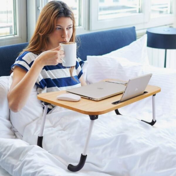 Woman using laptop table in bed with mug in hand.