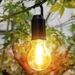 Hanging LED camping lantern with hook, casting warm light among tree leaves, creating a cozy outdoor atmosphere.