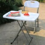 Folding coffee table set up outdoors with a mug, plate of food, and books, surrounded by greenery.