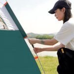 Woman adjusting the pole of an automatic pop-up camping tent in a grassy field, demonstrating ease of use.
