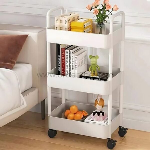 White 3-tier rolling organizer filled with books, decor, and fruit near a bed in a cozy bedroom.