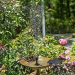 Solar-powered water fountain birdbath surrounded by blooming flowers and lush plants in a colorful garden.