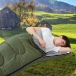 Man sleeping in a green portable sleeping bag on grassy terrain with scenic trees and barn backdrop.