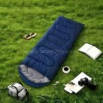Blue portable sleeping bag on grassy surface surrounded by travel gear—book, backpack, coffee, and camera.