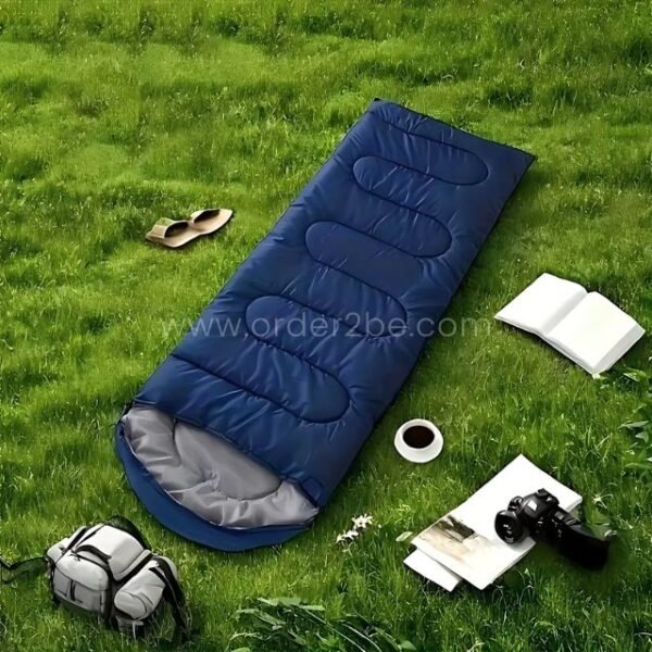Blue portable sleeping bag on grassy surface surrounded by travel gear—book, backpack, coffee, and camera.