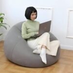 Woman working on a laptop while sitting on a gray inflatable bean bag chair for indoor use.