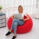 Woman sitting on a red inflatable bean bag chair for indoor use while working on a laptop in a bright room.